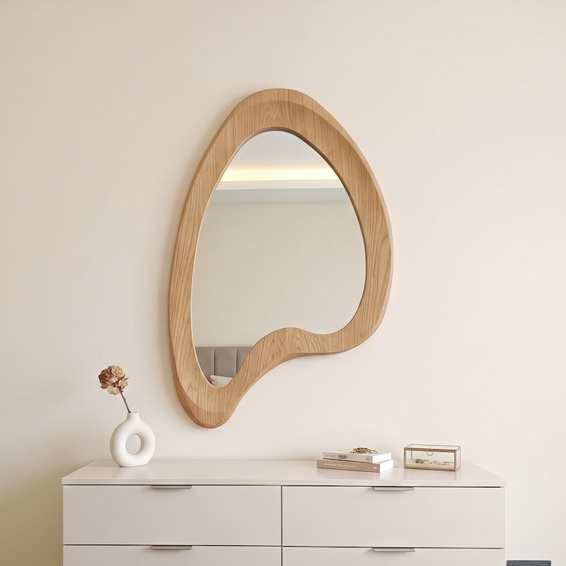May include: A wall-mounted mirror with an irregular, organic, amoeba-like shape and a light-toned wood grain frame. The mirror features a smooth, curved wooden border that surrounds the reflective glass surface, creating a distinct, asymmetrical silhouette.