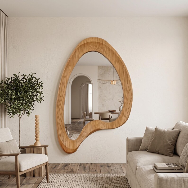 May include: A large wall mirror with an irregular, organic, amoeba-like shape and a light brown wood grain frame. The reflective surface shows a room interior with an arched doorway, a dining table, and chairs, creating a depth effect.