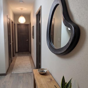 May include: An irregular, organic-shaped wall mirror with a dark, wood-grain frame mounted on a light-colored wall. The mirror features a thick, curved border that contrasts with the smooth, neutral surface of the wall behind it.