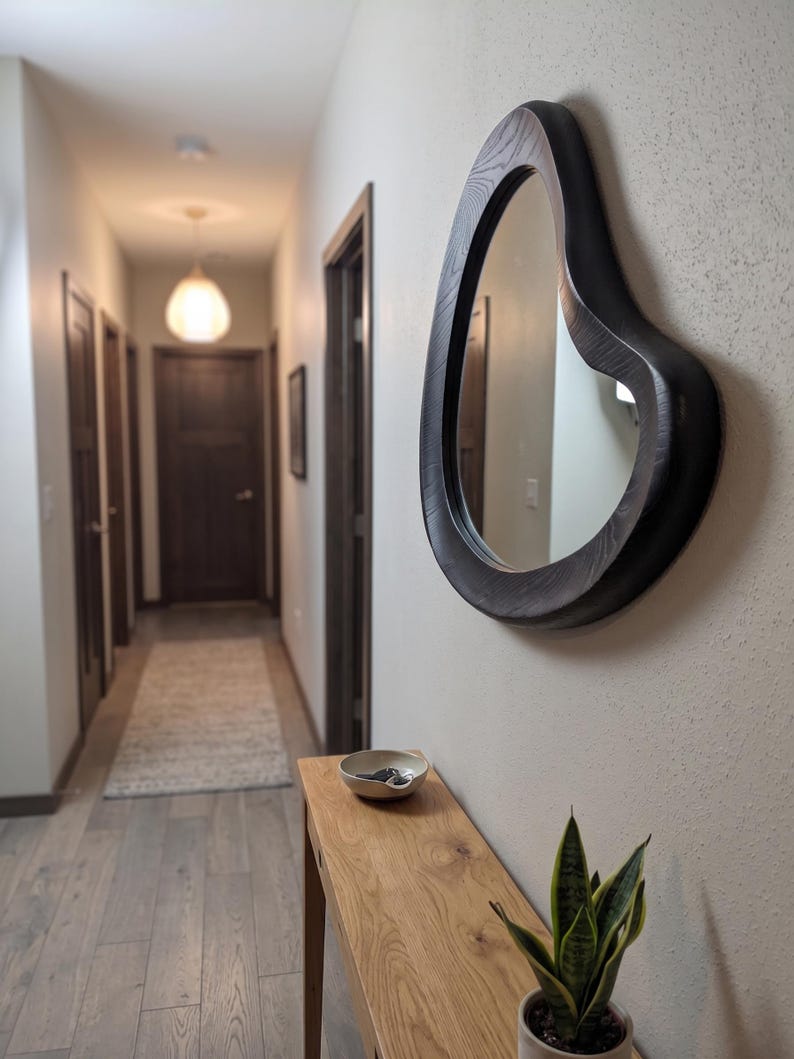 May include: An irregular, organic-shaped wall mirror with a dark, wood-grain frame mounted on a light-colored wall. The mirror features a thick, curved border that contrasts with the smooth, neutral surface of the wall behind it.