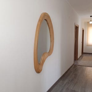 May include: A wall-mounted mirror featuring an asymmetrical, organic, wavy wooden frame with a light brown wood grain finish. The mirror has a smooth, curved shape that deviates from traditional geometric forms, providing a reflective surface.