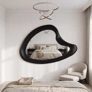May include: A large, wall-mounted mirror featuring an irregular, organic, amoeba-like shape with a thick, dark black frame. The reflective surface displays a bedroom interior, including a bed with neutral-toned bedding and two nightstands.
