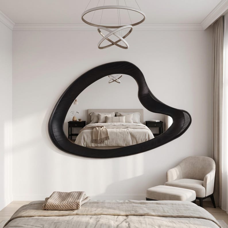 May include: A large, wall-mounted mirror featuring an irregular, organic, amoeba-like shape with a thick, dark black frame. The reflective surface displays a bedroom interior, including a bed with neutral-toned bedding and two nightstands.