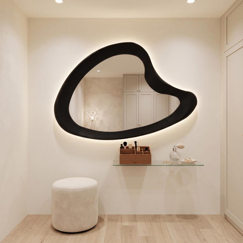 May include: An irregular, organic-shaped wall mirror with a thick black frame and integrated backlighting. The mirror is mounted above a clear glass floating shelf holding a brown organizer with makeup brushes and a small glass perfume bottle.