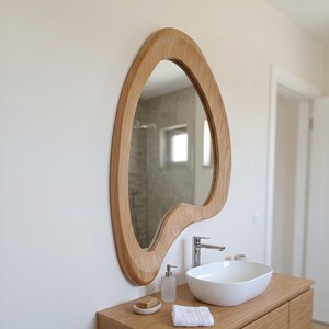 May include: A wall-mounted mirror with an irregular, organic, wavy wooden frame featuring a light brown wood grain pattern. The mirror is positioned above a white vessel sink, a silver faucet, a soap dispenser, and a small white towel.