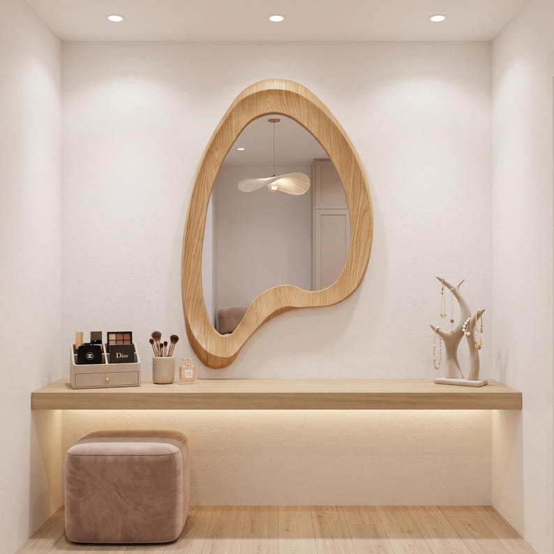 May include: A wall-mounted vanity area featuring a light wood floating shelf, a light wood organic-shaped mirror, a beige velvet ottoman, a makeup organizer with cosmetics, a brush holder, and a light-colored branch-style jewelry stand.