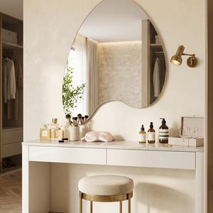 May include: A white vanity table with two drawers and a marble top, paired with a round beige upholstered stool on a gold metal base. Above the table hangs an asymmetrical, organic-shaped wall mirror, accompanied by a small gold wall-mounted lamp.