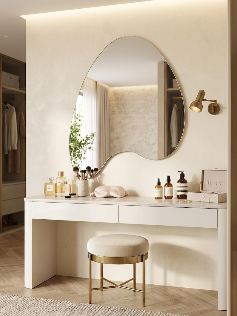 May include: A white vanity table with two drawers and a marble top, paired with a round beige upholstered stool on a gold metal base. Above the table hangs an asymmetrical, organic-shaped wall mirror, accompanied by a small gold wall-mounted lamp.