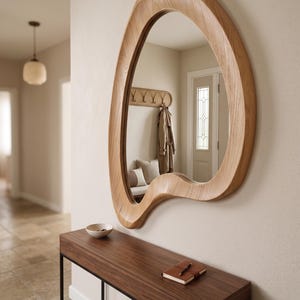 May include: A wall-mounted mirror featuring an irregular, organic, amoeba-like wooden frame with a light oak finish. The mirror has a smooth, curved border that creates a non-symmetrical shape, reflecting the interior space and a coat rack.