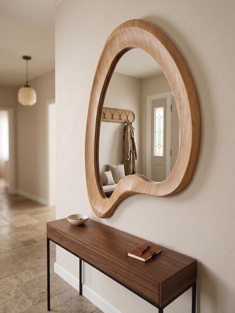 May include: A wall-mounted mirror featuring an irregular, organic, amoeba-like wooden frame with a light oak finish. The mirror has a smooth, curved border that creates a non-symmetrical shape, reflecting the interior space and a coat rack.