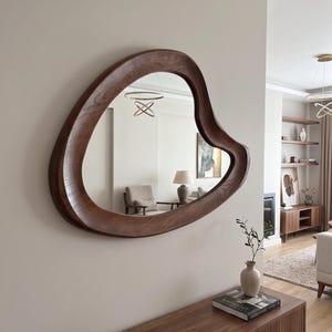 May include: A wall-mounted mirror featuring an irregular, organic, amoeba-like shape with a thick, dark brown wooden frame showing a visible wood grain texture. The mirror reflects a room interior with a lamp, chair, and ceiling light fixture.