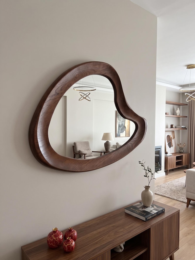 May include: A wall-mounted mirror featuring an irregular, organic, amoeba-like shape with a thick, dark brown wooden frame showing a visible wood grain texture. The mirror reflects a room interior with a lamp, chair, and ceiling light fixture.