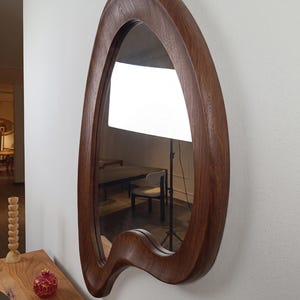 May include: A wall-mounted mirror featuring a thick, dark brown wooden frame with a distinct, asymmetrical, organic, and fluid shape. The wood grain is visible throughout the frame, which curves inward and outward, creating a unique, non-geometric silhouette.