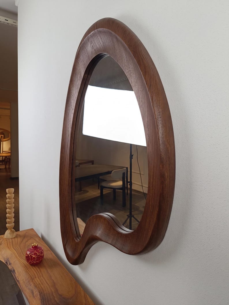 May include: A wall-mounted mirror featuring a thick, dark brown wooden frame with a distinct, asymmetrical, organic, and fluid shape. The wood grain is visible throughout the frame, which curves inward and outward, creating a unique, non-geometric silhouette.