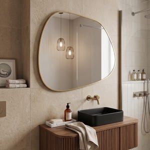 May include: An asymmetrical wall mirror with a thin gold frame, reflecting two hanging pendant lights. The mirror is mounted above a dark wood vanity featuring a black rectangular sink, a brass faucet, and a white marble tray with soap.