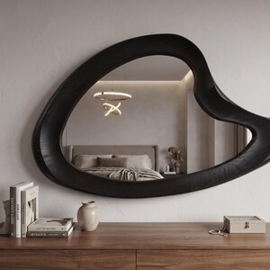 May include: A wall-mounted mirror featuring an irregular, organic, amoeba-like shape with a thick, dark black wooden frame that displays a visible wood grain texture. The reflective surface shows a bedroom interior with a bed and a light fixture.