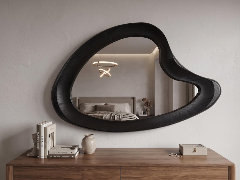 May include: A wall-mounted mirror featuring an irregular, organic, amoeba-like shape with a thick, dark black wooden frame that displays a visible wood grain texture. The reflective surface shows a bedroom interior with a bed and a light fixture.