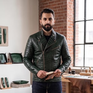 May include: A dark green leather jacket with a textured crocodile skin pattern, featuring a diagonal front zip, a stand-up collar with a snap button, and two zipped side pockets. The jacket is worn over a plain black shirt and dark blue jeans.