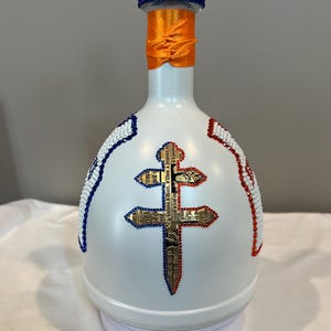 May include: A white bottle decorated with a central Lorraine cross made of gold-toned paper with French text, outlined in red rhinestones. The neck features an orange ribbon and two bands of red and blue rhinestones at the top rim.