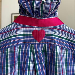 May include: A blue, green and white plaid shirt featuring a high ruffled collar, a bright pink ribbon trim around the neckline, and a solid pink embroidered heart centred on the back yoke above a box pleat detail in the fabric construction.