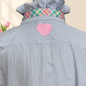 May include: A light blue and white pinstriped shirt featuring a ruffled collar with a multi-colored plaid trim. A pink embroidered heart patch is centered on the upper back of the shirt, positioned just below the collar and the plaid fabric band.