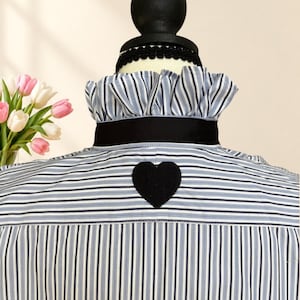 May include: A blue and white striped button-down shirt featuring a ruffled collar, a black ribbon trim around the neck, and a black heart-shaped patch centered on the upper back. The shirt is displayed on a mannequin torso.