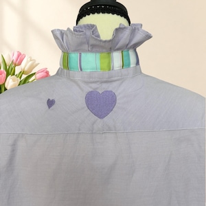 May include: A light purple and white micro-check shirt featuring a ruffled collar with a horizontal striped ribbon in shades of green, blue, and white. The back of the shirt is decorated with two embroidered purple hearts of different sizes.