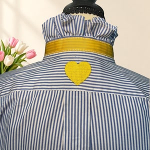 May include: A blue and white striped button-down shirt featuring a ruffled collar, a mustard yellow ribbon trim around the neck, and a yellow embroidered heart patch centered on the upper back yoke area of the garment.
