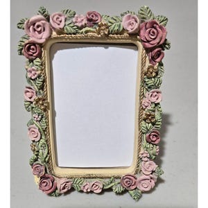 May include: A rectangular picture frame featuring a cream-colored inner border with a rope-like texture. The frame is decorated with sculpted green leaves, various shades of pink roses, and small gold-toned flower accents surrounding the center.