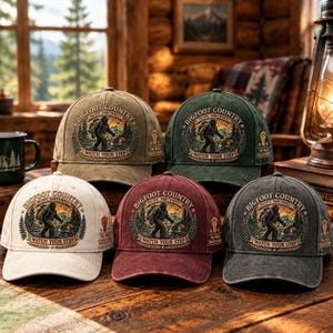 Può includere: Cinque cappellini da baseball in beige, verde, crema, bordeaux e grigio antracite. Ogni cappellino presenta una toppa circolare ricamata che raffigura la sagoma di un Bigfoot in un paesaggio boschivo con il testo Bigfoot Country e Watch Your Step.