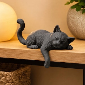 Könnte beinhalten: Eine dunkelgraue Katzenfigur in Schlafposition, deren Kopf auf den Vorderpfoten ruht und deren Schwanz nach oben gebogen ist. Die Katze befindet sich auf der Kante einer flachen Oberfläche, wobei ein Vorderbein über den Rand des Vorsprungs hängt.