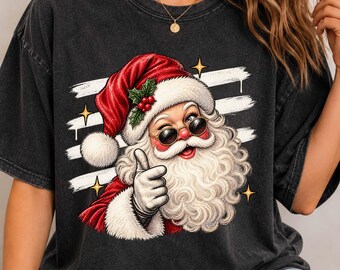 May include: A charcoal gray short-sleeve t-shirt featuring a graphic of Santa Claus wearing round sunglasses and a red hat with holly berries. Santa is giving a thumbs-up gesture against a background of three horizontal white brush strokes.