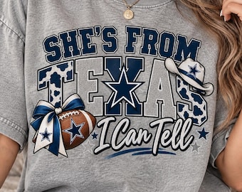 May include: A grey t-shirt featuring the text "SHE'S FROM TEXAS I Can Tell" in navy and white. The design includes a football with a blue and white bow, a cowboy hat, and a blue star graphic. The letters are filled with a blue and white cow print.