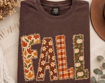 May include: A dark brown crew neck t-shirt featuring the word FALL in large, fabric-appliqued letters. Each letter has a distinct pattern: autumn leaves, white daisies on brown, orange plaid, and white pumpkins on green, all accented with rhinestones.