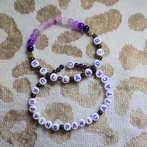 May include: Two beaded bracelets featuring purple and white beads, small gold star charms, and white letter beads. One bracelet spells out "HOLLANDER" and the other spells out "HAVING PANIC ATTACK" with a small purple heart bead in the center.