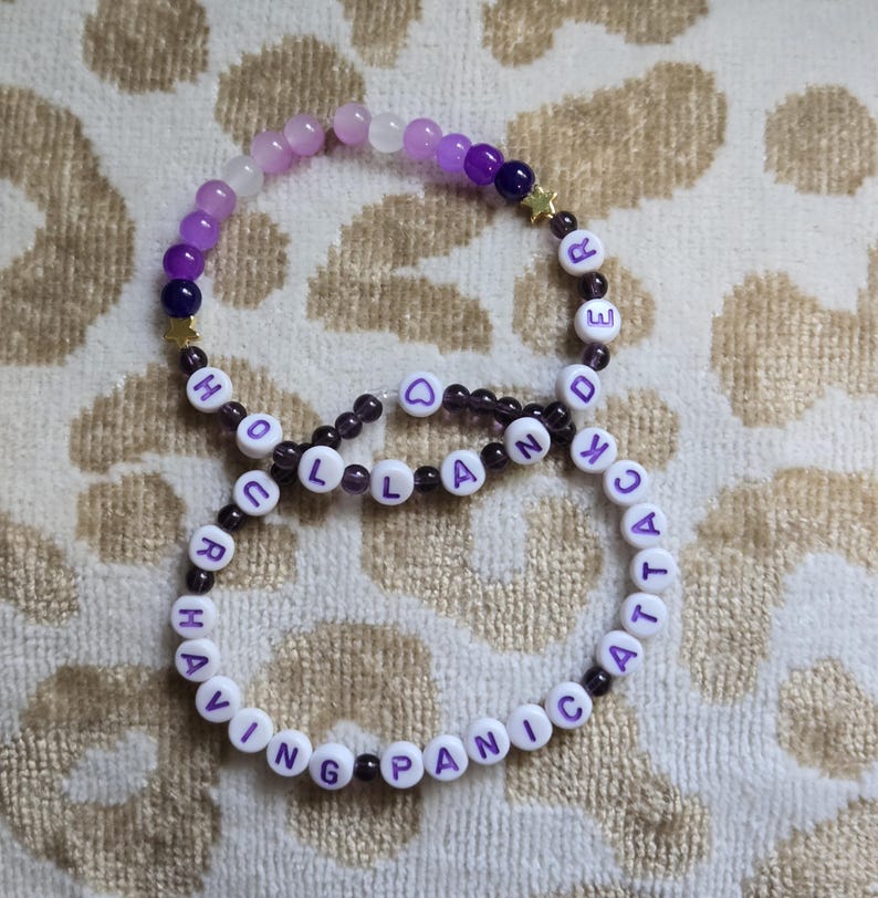 May include: Two beaded bracelets featuring purple and white beads, small gold star charms, and white letter beads. One bracelet spells out "HOLLANDER" and the other spells out "HAVING PANIC ATTACK" with a small purple heart bead in the center.