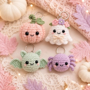 May include: Four crochet keychains featuring a pastel pink pumpkin with a green stem, a white ghost wearing a floral crown and bow, a light green bat with wings, and a light purple spider with eight legs, each with black bead eyes and pink cheeks.