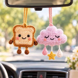 Può includere: Due ornamenti all'uncinetto da appendere allo specchietto retrovisore dell'auto. Uno è una fetta di pane tostato beige con un viso sorridente, crosta marrone e piccole braccia e gambe. L'altro è una nuvola rosa chiaro con un viso sorridente, gocce di pioggia rosa e una stella gialla.