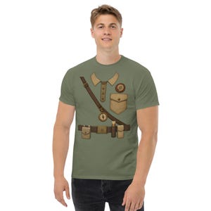 May include: An olive green t-shirt featuring a printed graphic of an explorer uniform. The design includes a tan collar, a chest pocket, a circular badge, a brown shoulder strap with a hanging compass, and a utility belt with pouches and tools.