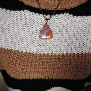 May include: A triangular pendant featuring a polished, multi-colored stone with swirls of red, orange, and white, suspended from a dark copper-toned metal chain. The pendant rests against a knit sweater with horizontal stripes in black, white, and tan.