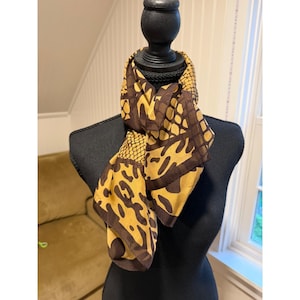 May include: A square scarf featuring a bold animal print pattern in shades of mustard yellow and dark brown. The design incorporates a mix of leopard spots and a textured snakeskin-like grid motif, tied neatly around the neck of a display form.