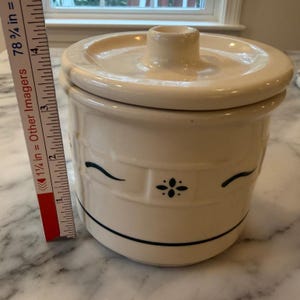 May include: A cream-coloured ceramic canister with a matching lid, featuring a dark blue embossed basket-weave pattern and a central floral motif. A vertical measuring tape placed beside the canister indicates a height of approximately 10 cm.