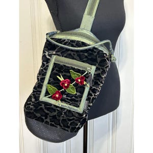 May include: A black and dark grey velvet sling bag with a leopard print pattern. The bag features a front zippered pocket adorned with a decorative branch of three red velvet flowers and green leaves. A small tag with the text GOODY is attached.