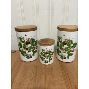 May include: A set of three white ceramic canisters of varying heights, each featuring a botanical strawberry pattern with green leaves, white flowers, and red berries. Each cylindrical container is topped with a round wooden lid.