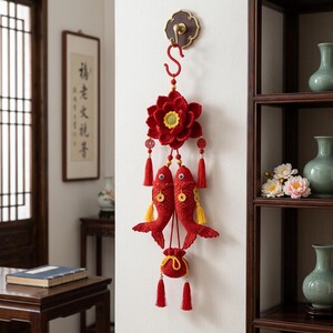 May include: A red crocheted wall hanging featuring a large lotus flower at the top, two fish figures with yellow tassels and gold-colored coins, and a small drawstring pouch at the bottom, all suspended from a red S-shaped hook on a wall.
