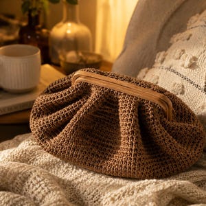 May include: A brown crochet clutch bag featuring a gathered, pleated texture and a light-colored wooden frame closure. The bag has a rounded, soft silhouette and is constructed from a woven fiber material with a consistent, dense stitch pattern.