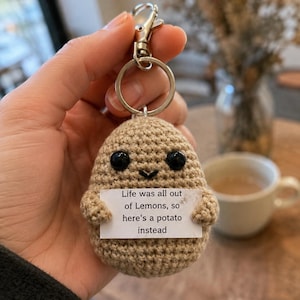 May include: A tan crocheted potato keychain with black plastic eyes and a small stitched smile. The potato holds a white rectangular sign that reads: Life was all out of Lemons, so here's a potato instead. It features a silver metal keyring.