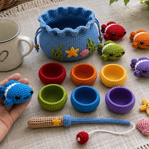 May include: A blue crocheted drawstring pouch with sea-themed appliques, six small colorful crocheted bowls, a crocheted fishing rod, and several small crocheted sea creatures including fish, a starfish, and a seashell, alongside a white mug.