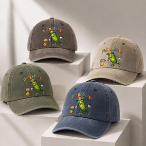 Puede incluir: Cuatro gorras de béisbol en gris carbón, beige, verde oliva y azul marino. Cada gorra presenta un gráfico de un pepinillo de dibujos animados jugando al pickleball con una pala y una pelota, con la palabra PICKLEBALL en letras coloridas.