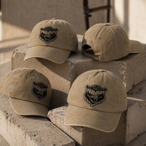 Puede incluir: Cuatro gorras de béisbol de color tostado hechas de algodón lavado. Cada gorra presenta un gráfico bordado en negro de una cara de oso estilizada en el panel frontal. Las gorras tienen visera curva, ojales de ventilación y hebilla metálica.
